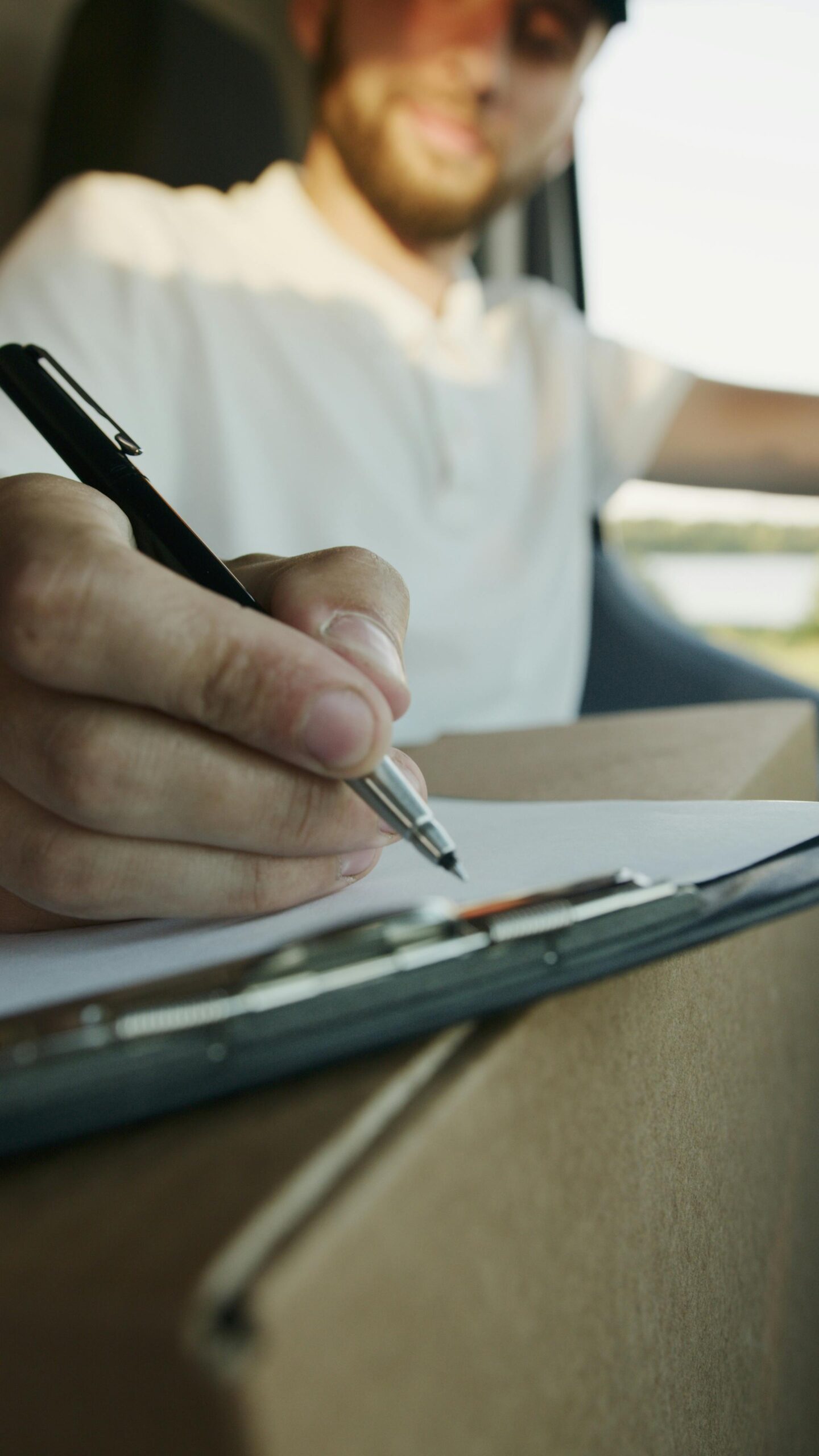 Close-up of a delivery driver writing on a clipboard while holding a package inside a vehicle.
