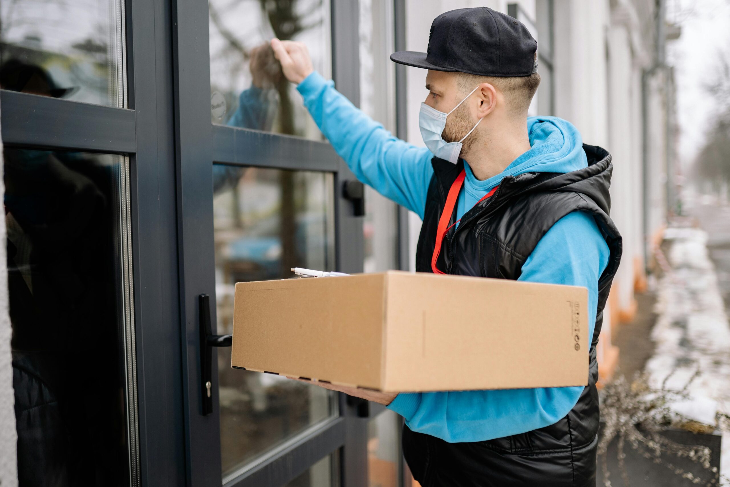 Male courier wearing a face mask delivers a package to a modern door, promoting contactless delivery.